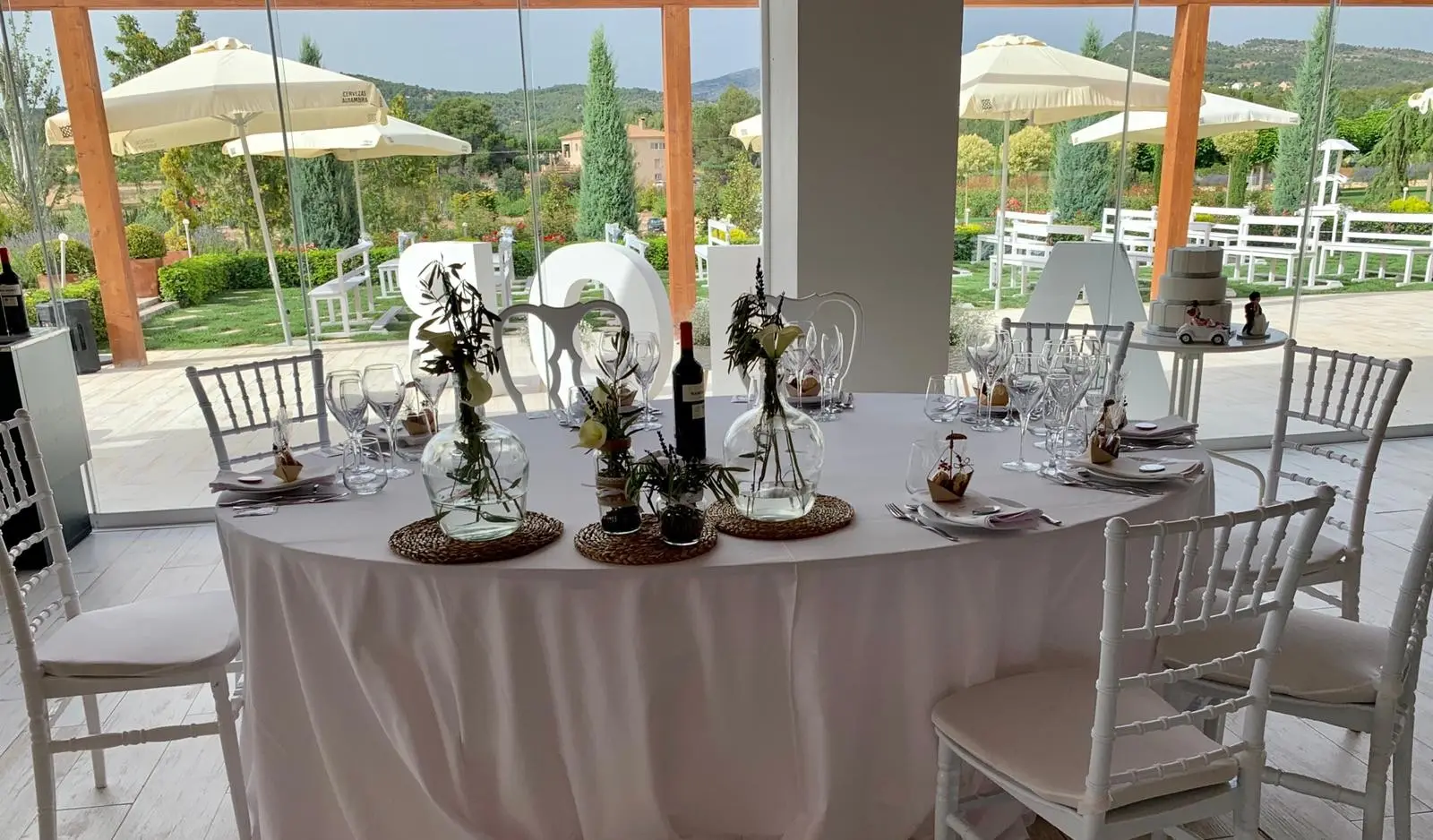 Una mesa redonda con mantel blanco y centro de flores del campo, sillas blancas y sombrillas junto a un ventanal con vistas a las montañas
