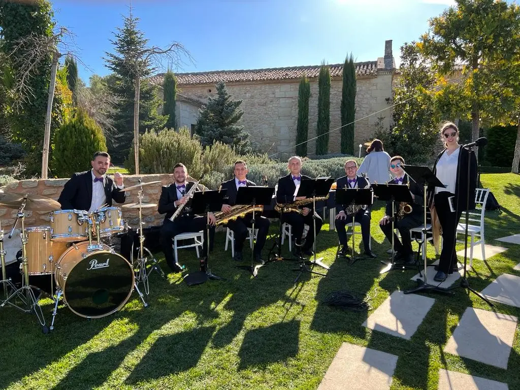 Un grupo de música con batería, saxofones y cantante, tocando en el césped bajo los pinos, ante un muro de piedra con cipreses
