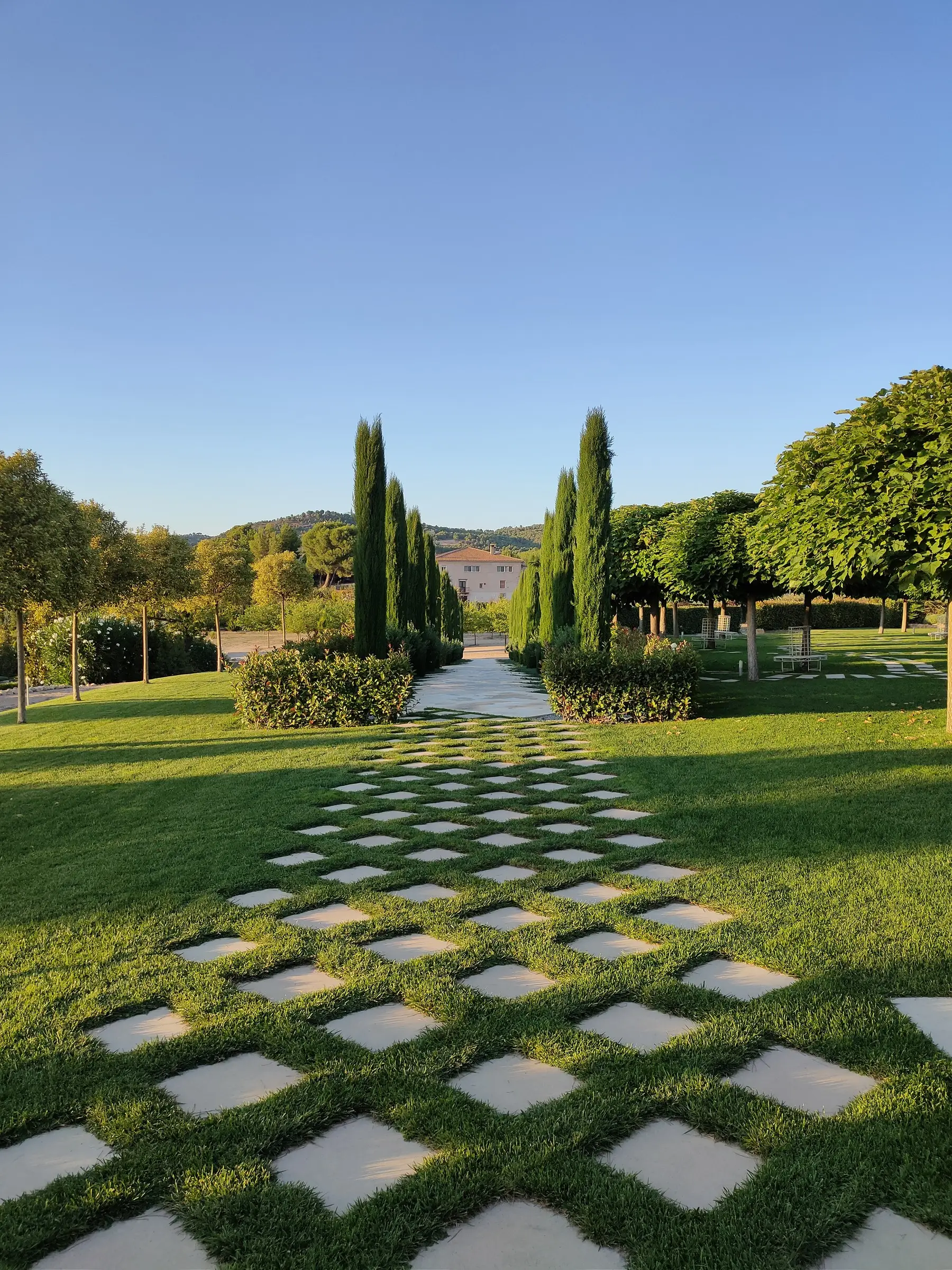 Un paseo de losas de piedra entre el césped, con cipreses altos a los lados y, al fondo, una casa de fachada clara con tejado de teja bajo el cielo del atardecer